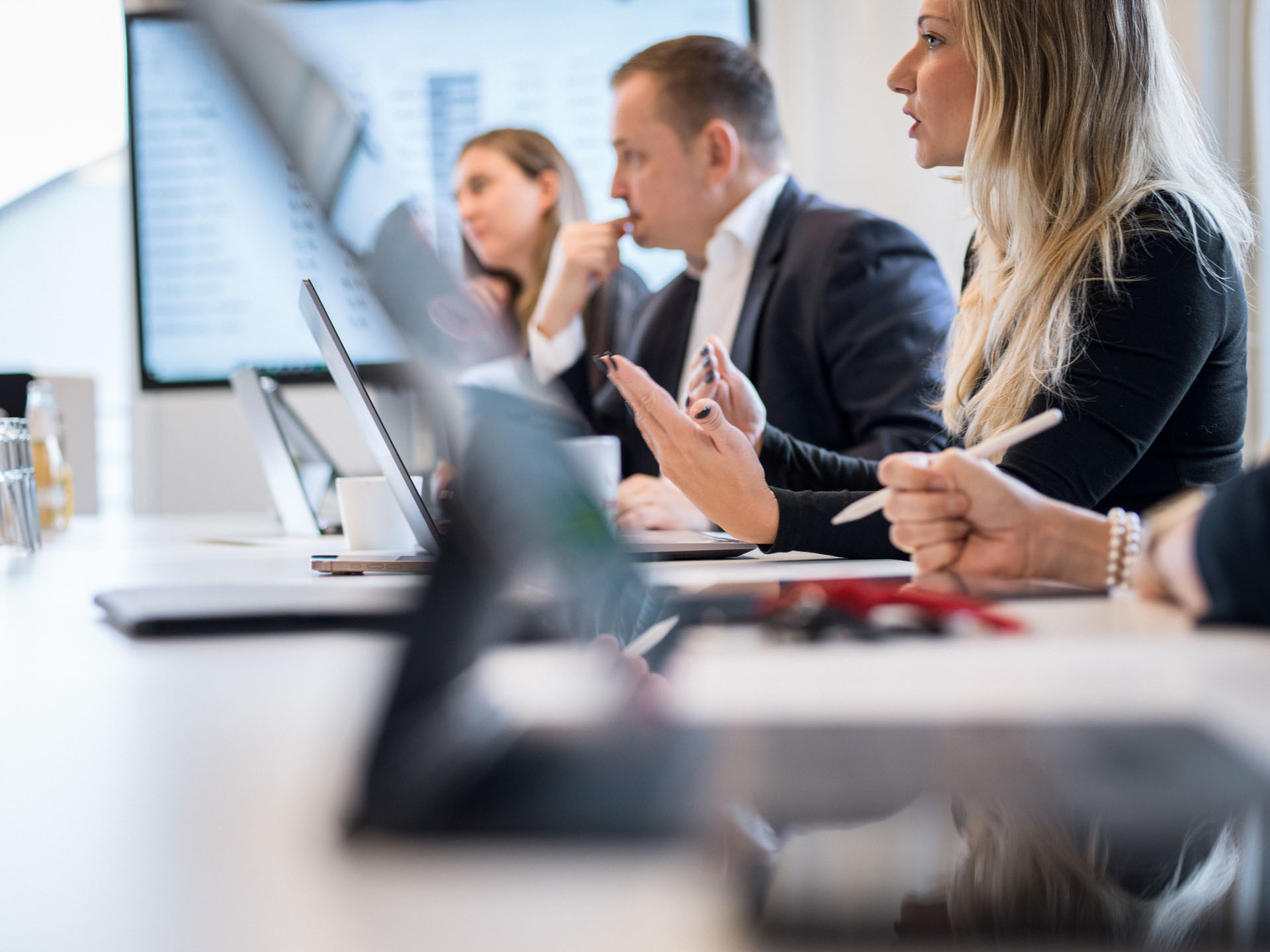 In einem modernen Besprechungsraum diskutieren mehrere Personen aktiv, während sie Laptops und Tablets nutzen. Im Vordergrund sind Hände mit einem Stift und Notizen zu sehen, während im Hintergrund eine Person am Tisch spricht. An der Wand ist ein Bildschirm mit Daten oder Diagrammen sichtbar.