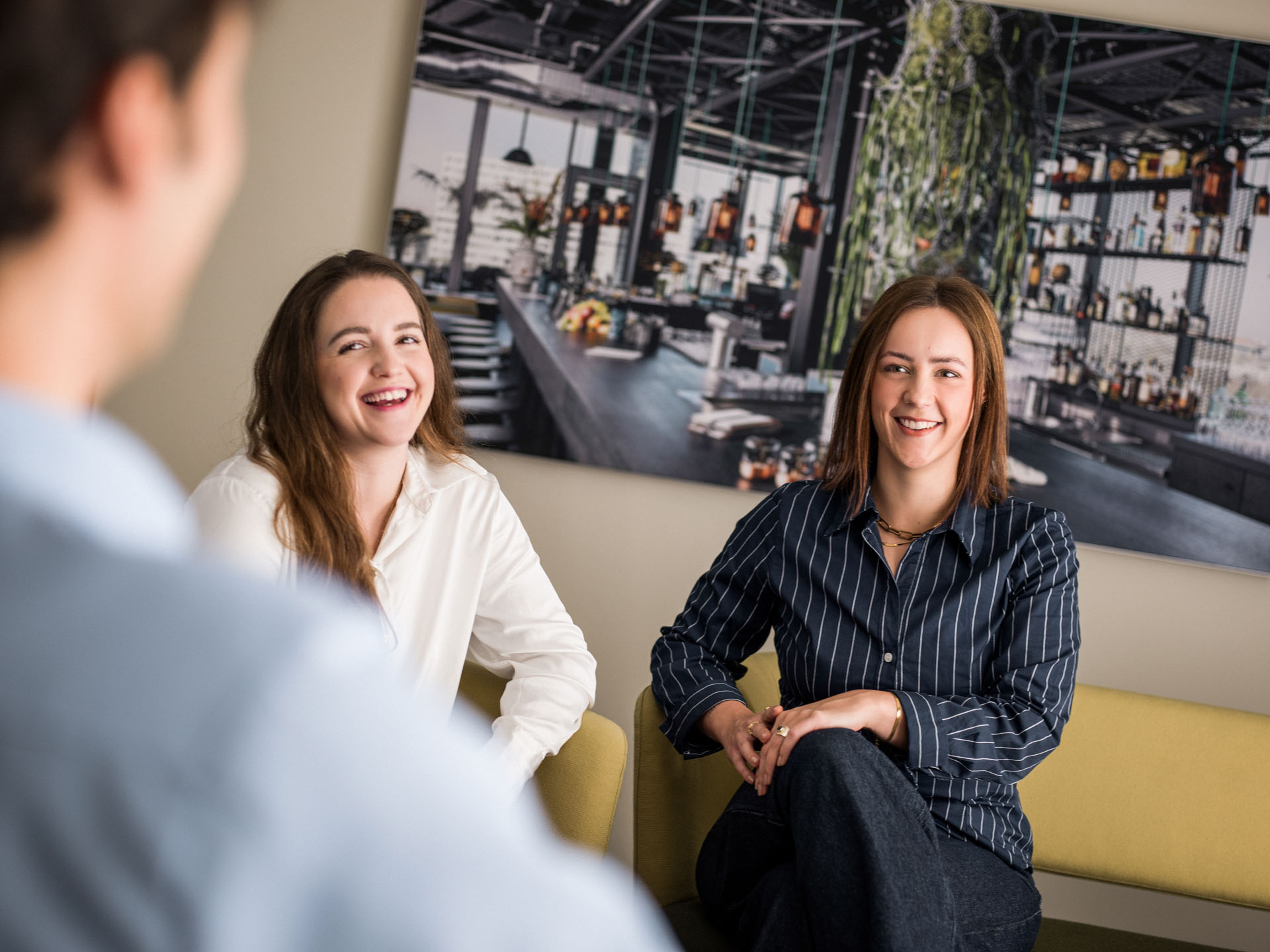 Zwei Frauen sitzen auf einem Sofa und lächeln, während sie mit einer anderen Person sprechen. Im Hintergrund ist ein großes Bild einer modernen Bar oder Lounge zu sehen.