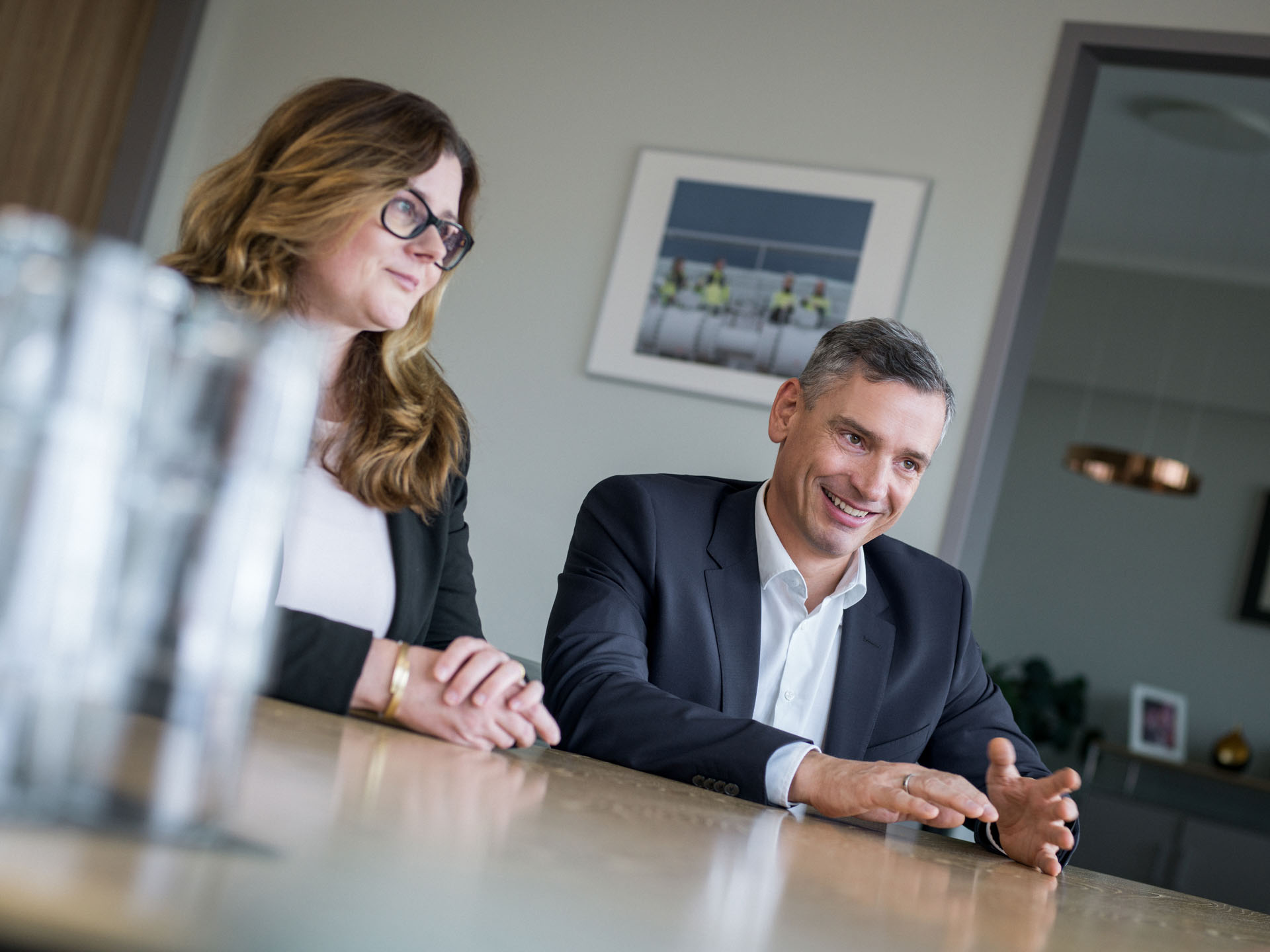 Zwei Personen sitzen an einem Tisch in einem modernen Konferenzraum; der Mann in Anzug gestikuliert und lächelt, während die Frau neben ihm aufmerksam zuhört. Im Hintergrund sind organisatorische Details sichtbar, inklusive einer Wanddekoration.