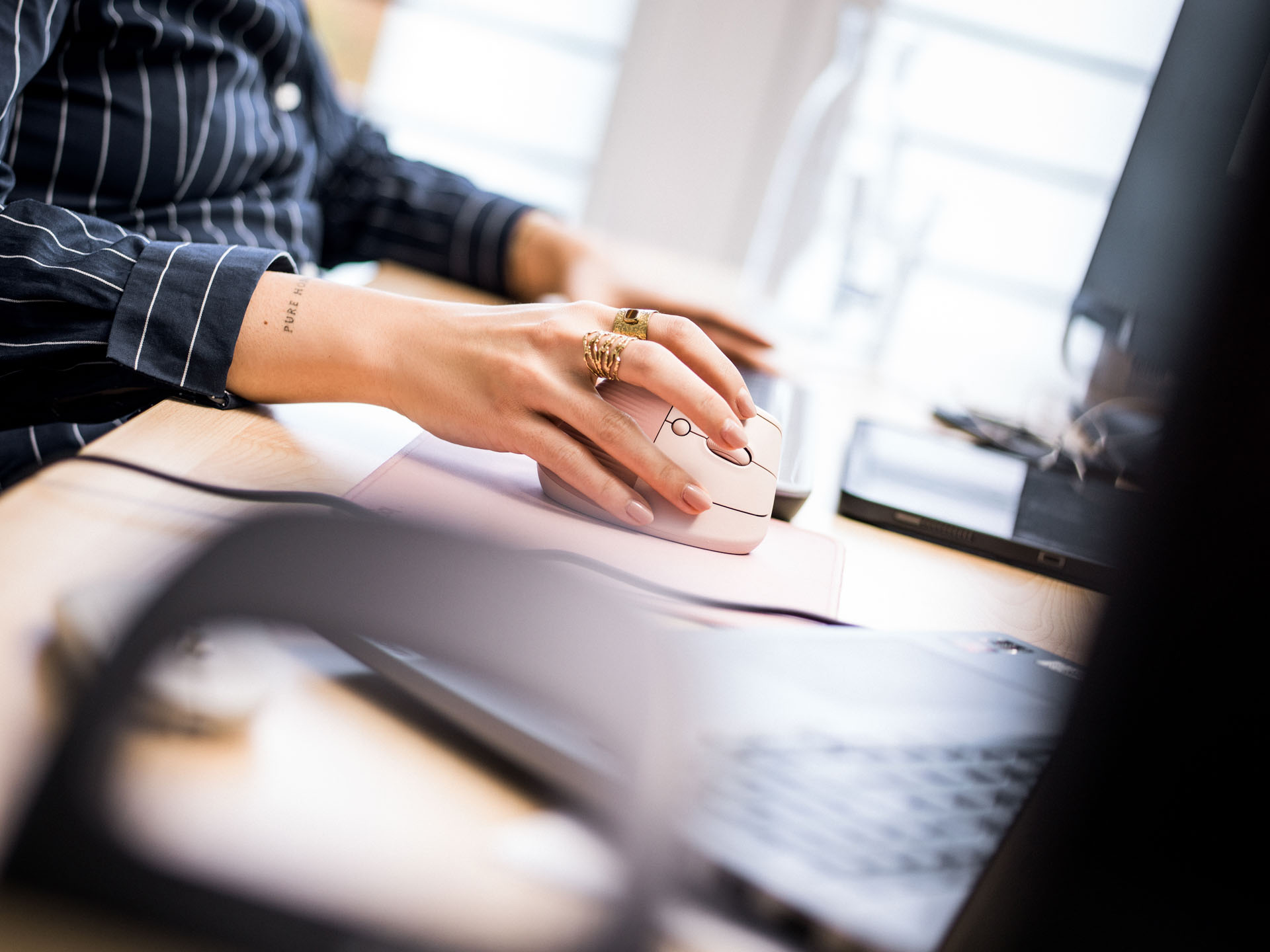 Eine Hand mit einem Ring bewegt eine Computermaus über ein Mauspad, während auf einem Schreibtisch ein Laptop und andere Bürogegenstände zu sehen sind.