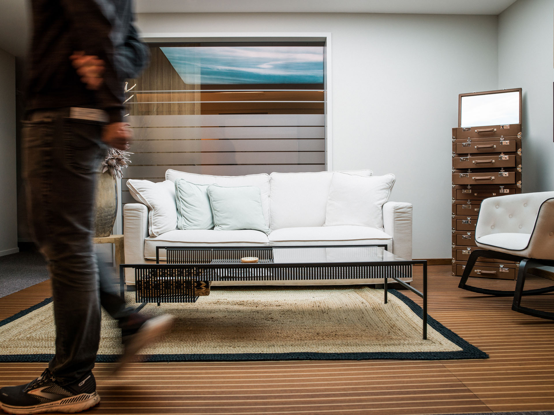 Ein moderner Wohnraum mit einem weißen Sofa, zwei Kissen, einem niedrigen Tisch und einem Teppich. Im Hintergrund sind Holzpaneele und eine Wand mit einem großformatigen Bild sichtbar. Eine Person in Jeans und jackets bewegt sich im Vordergrund.