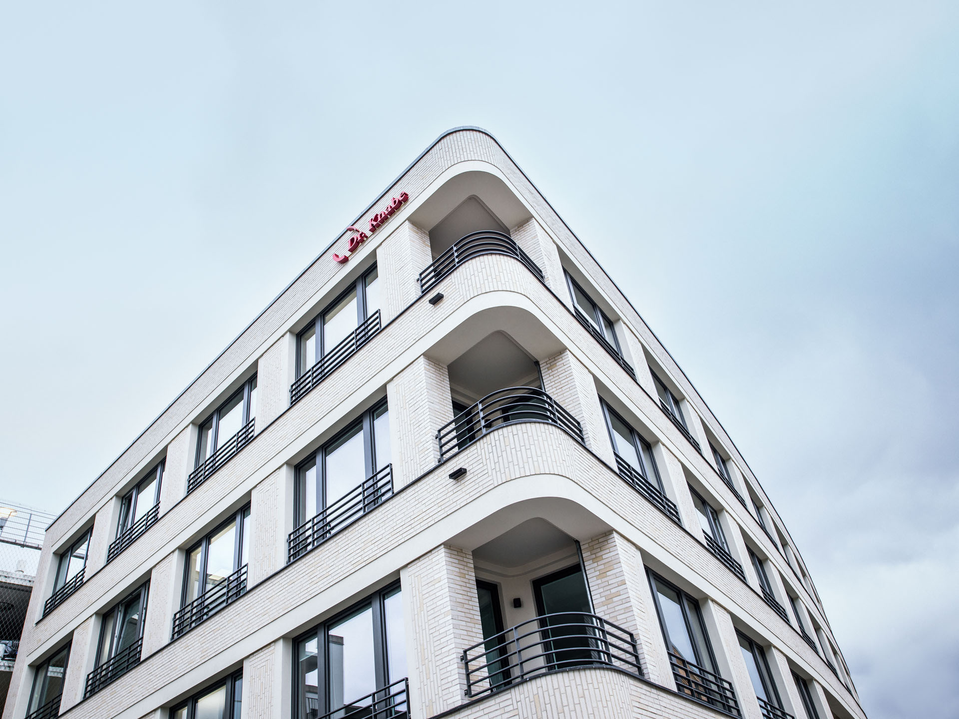 Das Bild zeigt die Fassade eines modernen, mehrstöckigen Gebäudes mit weißer Ziegelverkleidung und großen Fenstern, das einen gewölbten oberen Rand hat. Auf der Fassade ist der Schriftzug "La Maison" sichtbar. Der Himmel ist bewölkt.