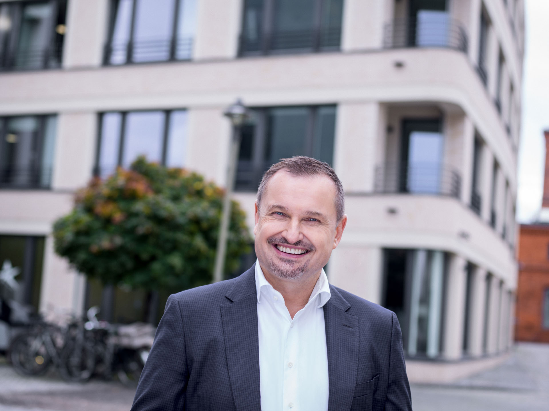 Steuerberater Potsdam Prof. Knabe - Ein lächelnder Mann in einem dunkelgrauen Anzug steht vor einem modernen, mehrstöckigen Gebäude mit großen Fenstern und einem grünen Baum im Hintergrund.