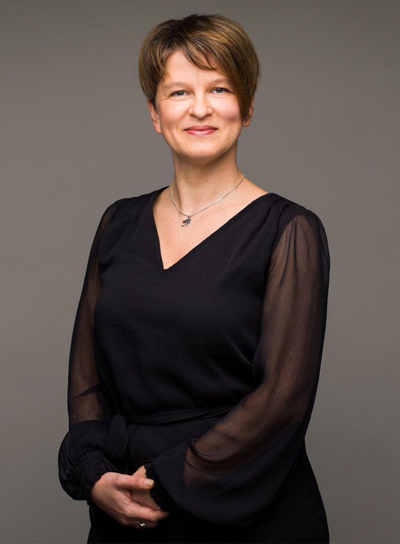Eine lächelnde Frau mit kurzen, braunen Haaren trägt ein schwarzes Kleid mit langen, transparenten Ärmeln und steht vor einem neutralen Hintergrund.