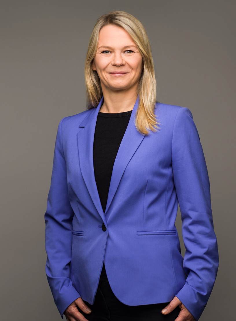 Eine lächelnde Frau mit langen, blonden Haaren trägt einen blauen Blazer und steht vor einem grauen Hintergrund.