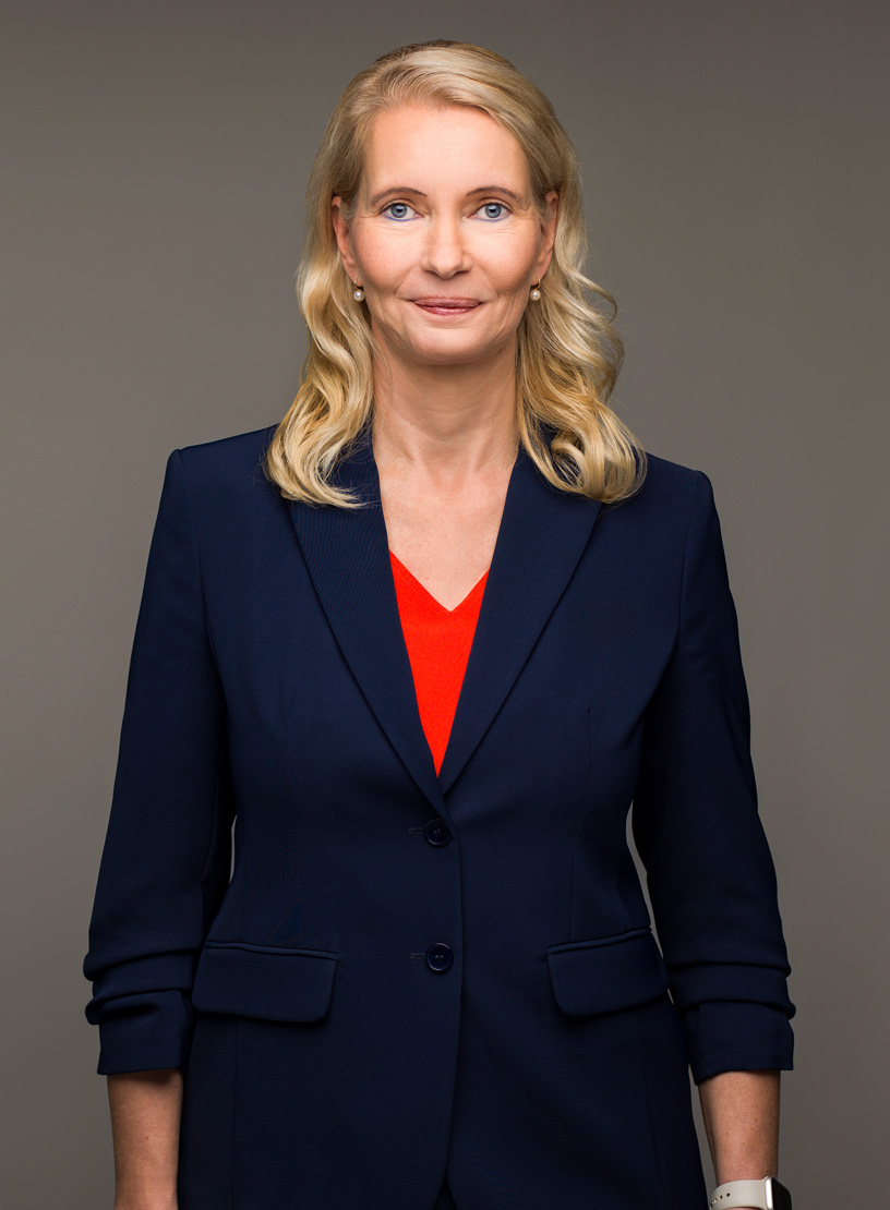 Eine lächelnde Frau mit langen, blonden Haaren trägt einen dunkelblauen Blazer über einem roten Oberteil. Sie steht vor einem grauen Hintergrund und blickt direkt in die Kamera.