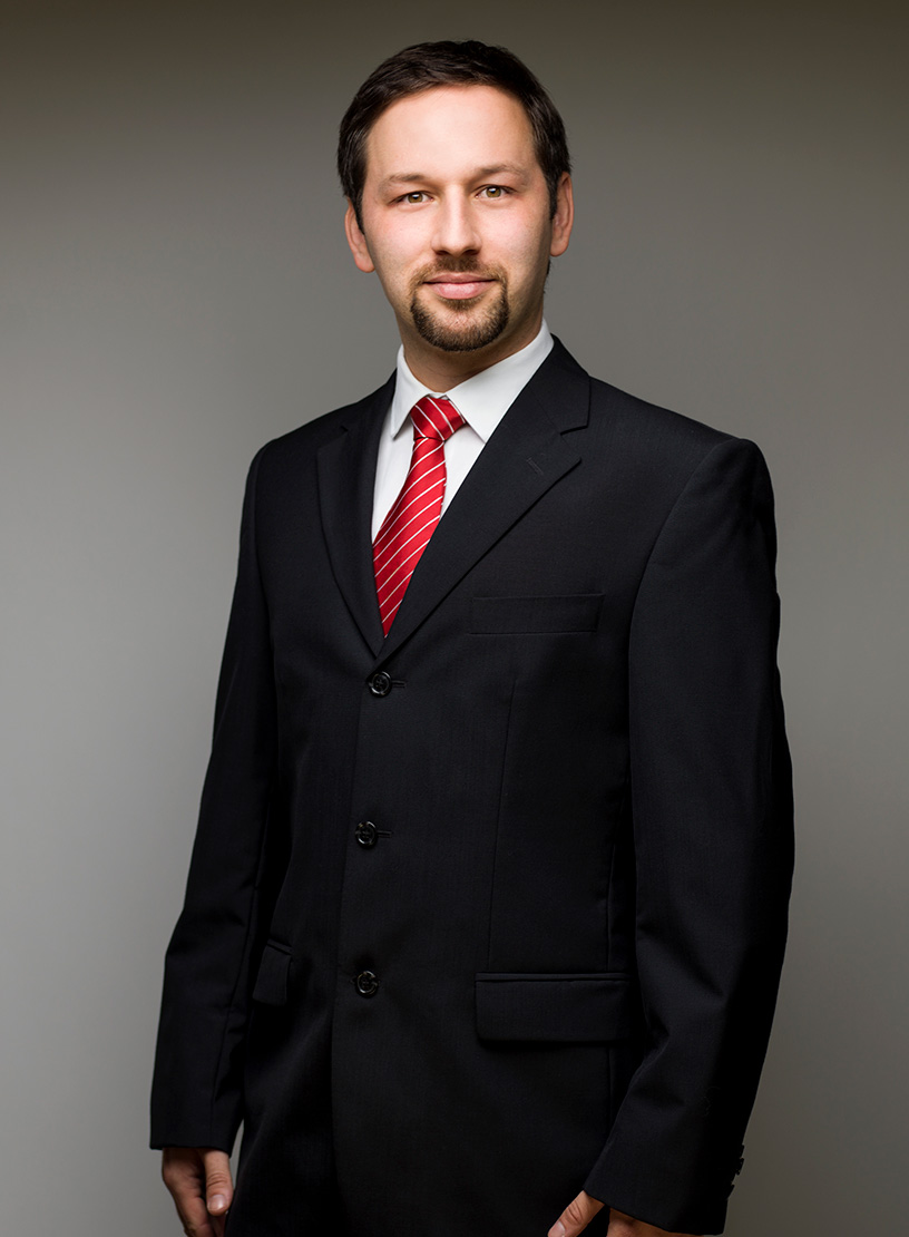 Ein Mann in einem schwarzen Anzug mit einem weißen Hemd und einer roten Krawatte steht vor einem neutralen Hintergrund und schaut direkt in die Kamera.