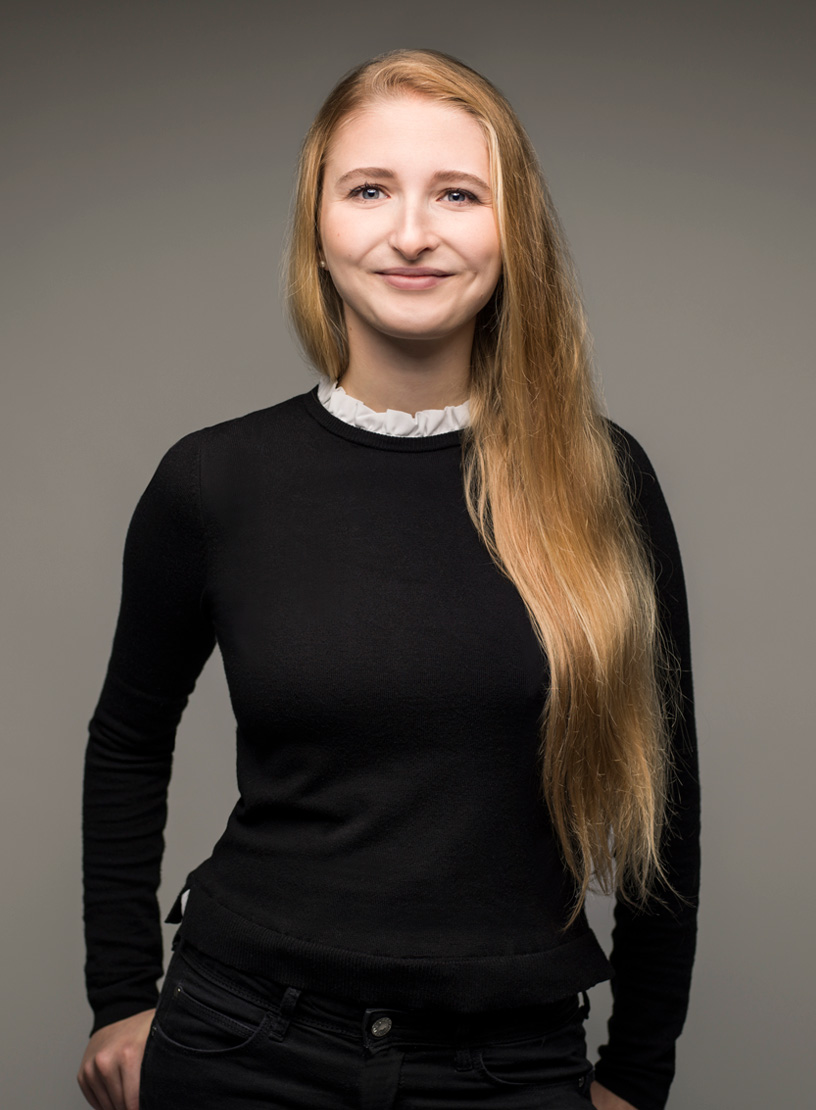Eine Person mit langen, blonden Haaren lächelt freundlich in die Kamera, dressed in einem schwarzen Pullover mit einem weißen Kragen. Der Hintergrund ist einfarbig grau.