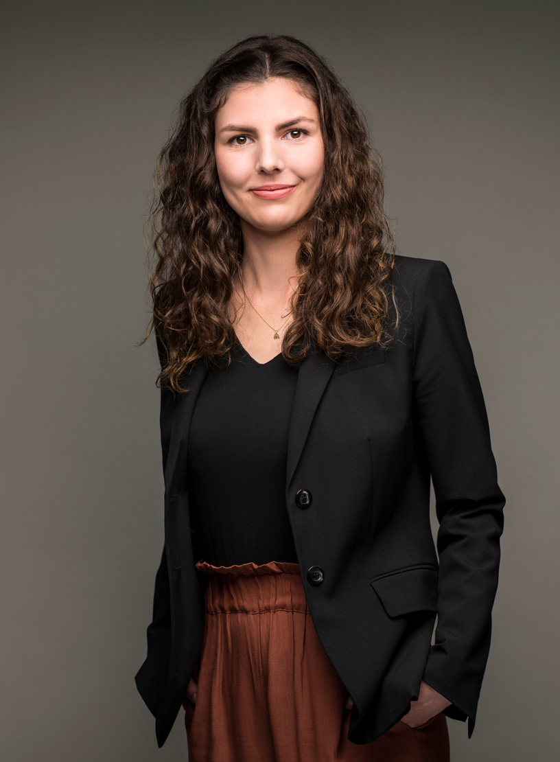 Eine Frau mit lockigem, braunem Haar trägt einen schwarzen Blazer und eine schwarze Bluse sowie eine rustikale, braune Hose. Sie lächelt freundlich und steht vor einem neutralen Hintergrund.