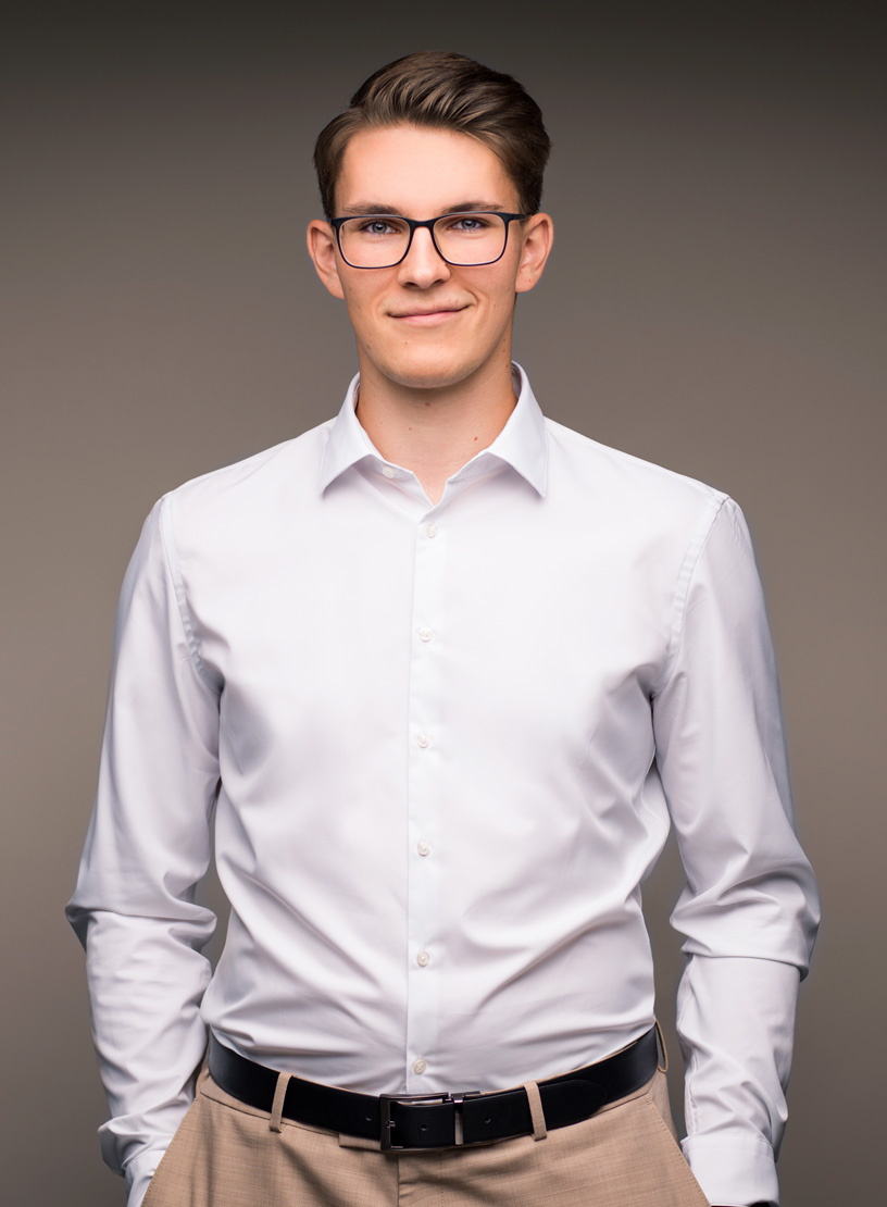 Ein junger Mann mit Brille trägt ein weißes Hemd und eine beige Hose. Er steht lächelnd vor einem neutralen, grauen Hintergrund.