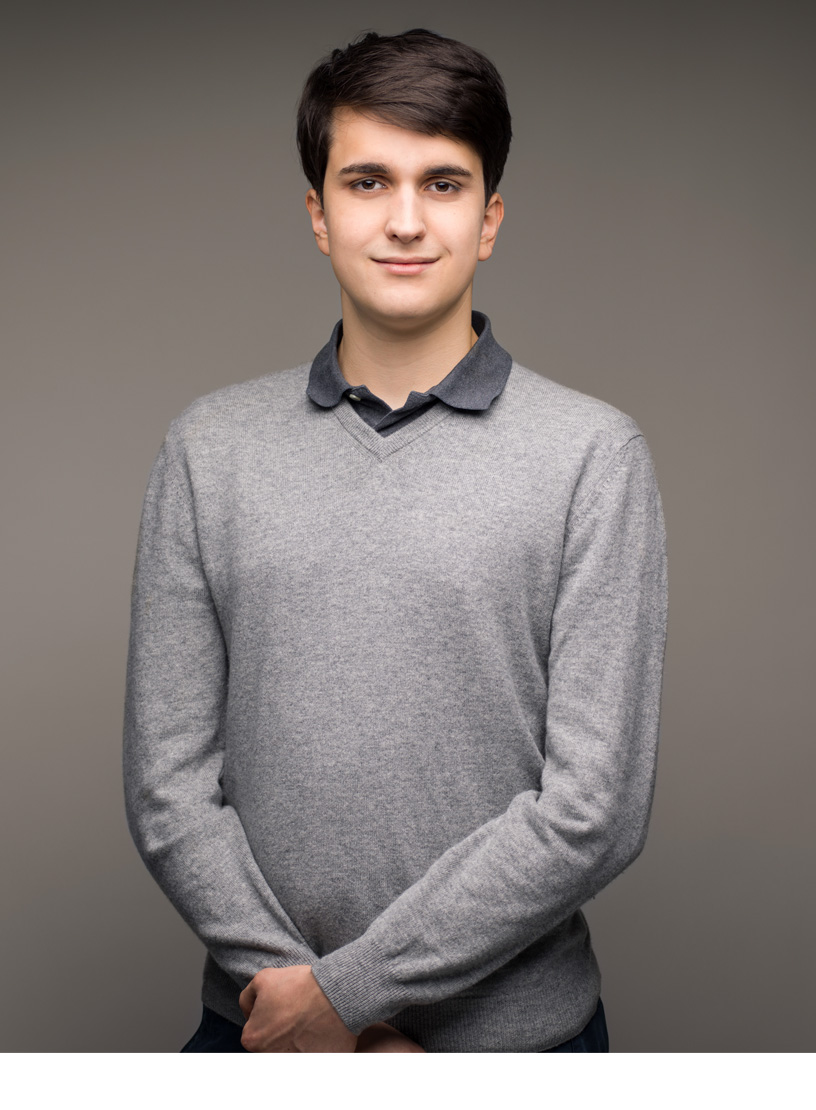 Ein junger Mann mit dunklen Haaren steht mit verschränkten Händen vor einem neutralen, grauen Hintergrund und trägt einen grauen Pullover über einem blauen Poloshirt.