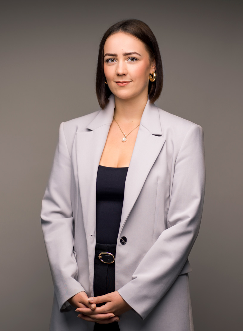 Eine Person in einem grauen Blazer und schwarzem Oberteil steht mit verschränkten Armen vor einem neutralen Hintergrund. Sie hat kurzes, braunes Haar und trägt goldene Ohrringe.