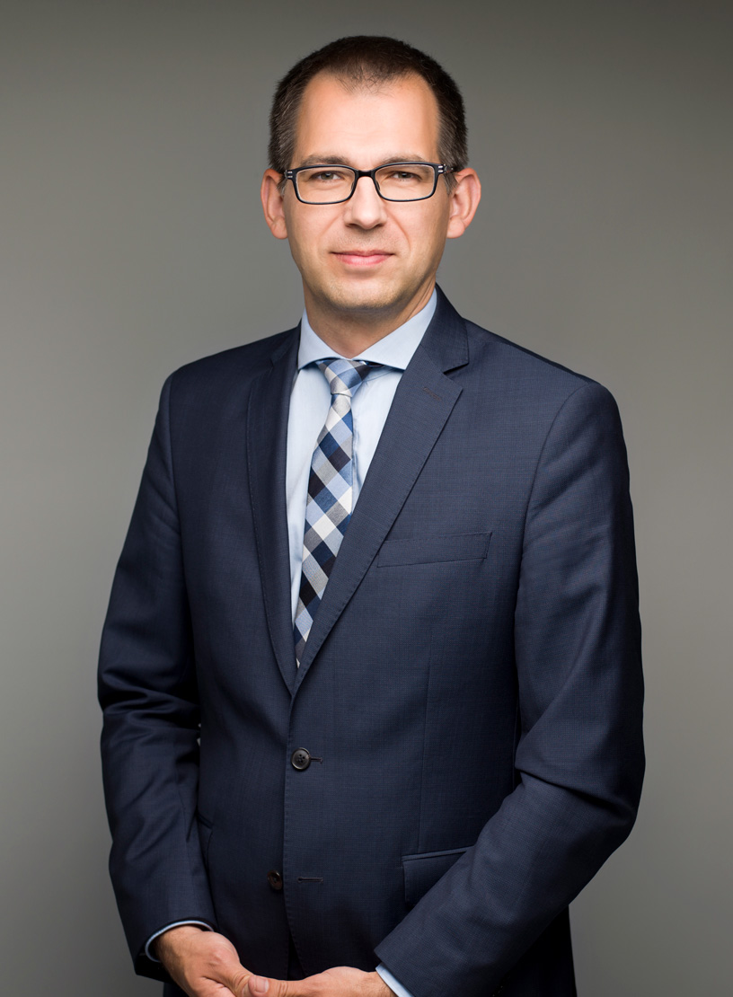 Eine geschäftlich gekleidete Person mit Brille steht aufrecht und hält die Hände vor sich, während sie in die Kamera schaut. Der Hintergrund ist neutral und glatt. Die Person trägt einen dunkelblauen Anzug und ein kariertes Hemd.