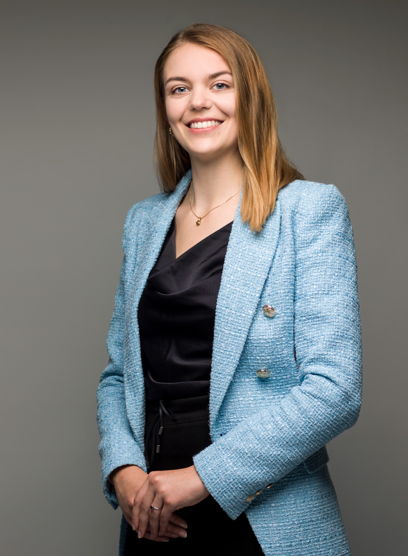 Eine smilende Frau mit langen, blonden Haaren trägt eine hellblaue Jacke über einem schwarzen Kleid und steht vor einem neutralen Hintergrund.