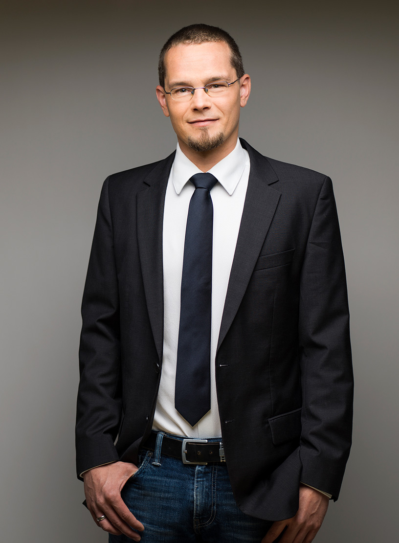 Ein Mann in einem Anzug mit einem weißen Hemd und einer dunklen Krawatte steht lässig mit einer Hand in der Hosentasche vor einem neutralen Hintergrund.