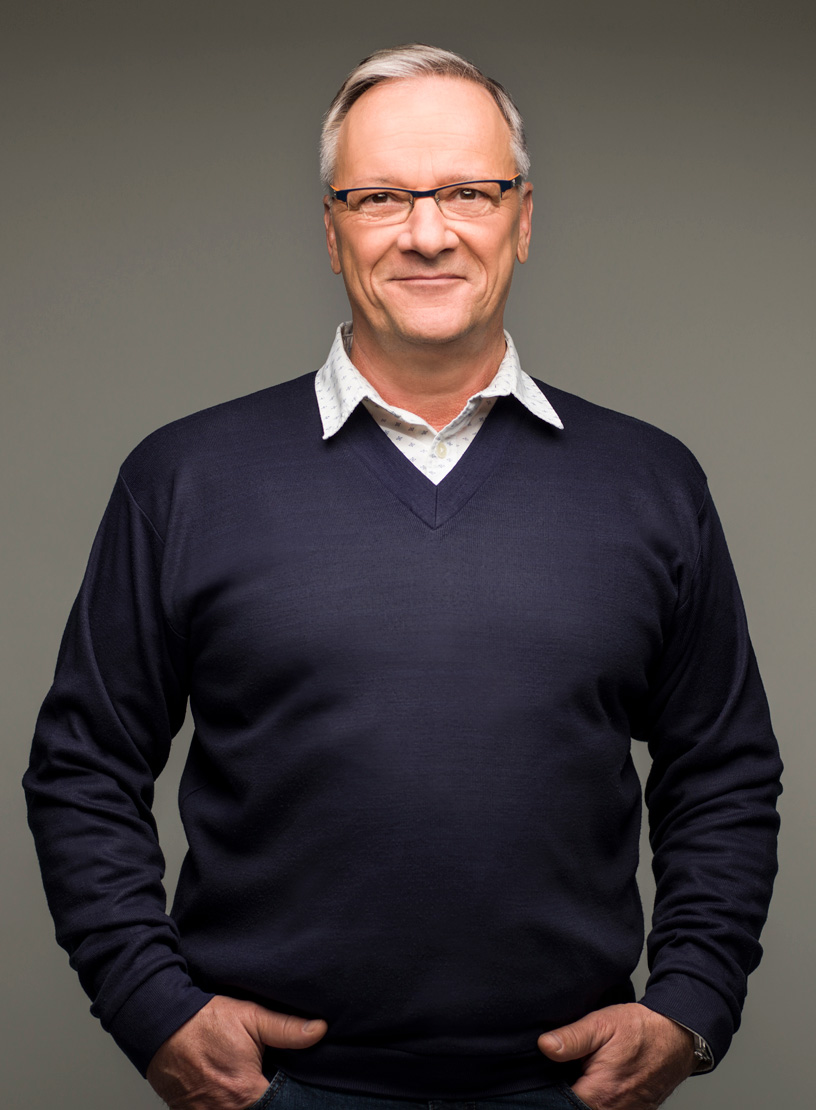 Ein älterer Mann mit Brille trägt einen dunkelblauen Pullover über einem hellen Hemd und steht mit verschränkten Händen vor einem einfarbigen Hintergrund.