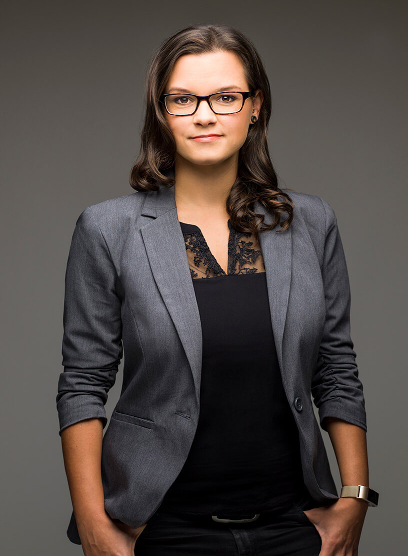 Eine selbstbewusste Frau mit schulterlangen, braunen Haaren und Brille trägt ein graues Blazer-Jackett über einem schwarzen Oberteil. Sie steht vor einem neutralen Hintergrund und schaut direkt in die Kamera.