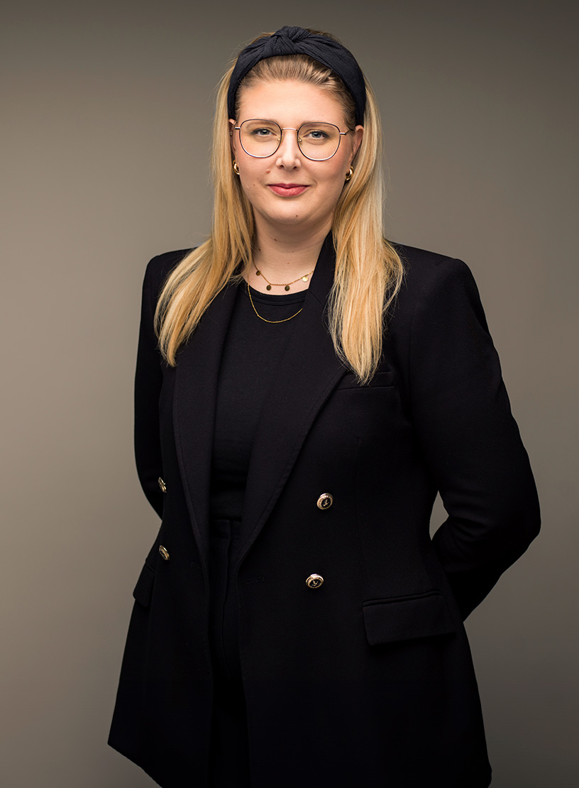 Eine Frau mit langen blonden Haaren und einer schwarzen Bluse trägt einen eleganten schwarzen Blazer. Sie hat eine modische Brille auf und steht mit verschränkten Händen vor einem neutralen Hintergrund.