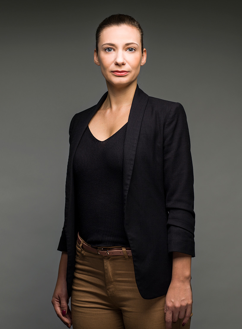 Eine Frau mit langen, glatten Haaren steht vor einem grauen Hintergrund und trägt einen schwarzen Blazer über einem schwarzen Oberteil sowie braunen Hosen. Sie schaut direkt in die Kamera und hat einen neutralen Gesichtsausdruck.