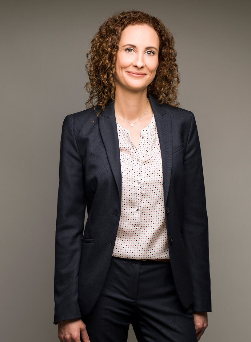 Eine Frau mit lockigem, braunem Haar trägt einen dunkelblauen Blazer über einer blassbeigen Bluse mit kleinen Punkten und lächelt in die Kamera. Der Hintergrund ist neutral und hell.