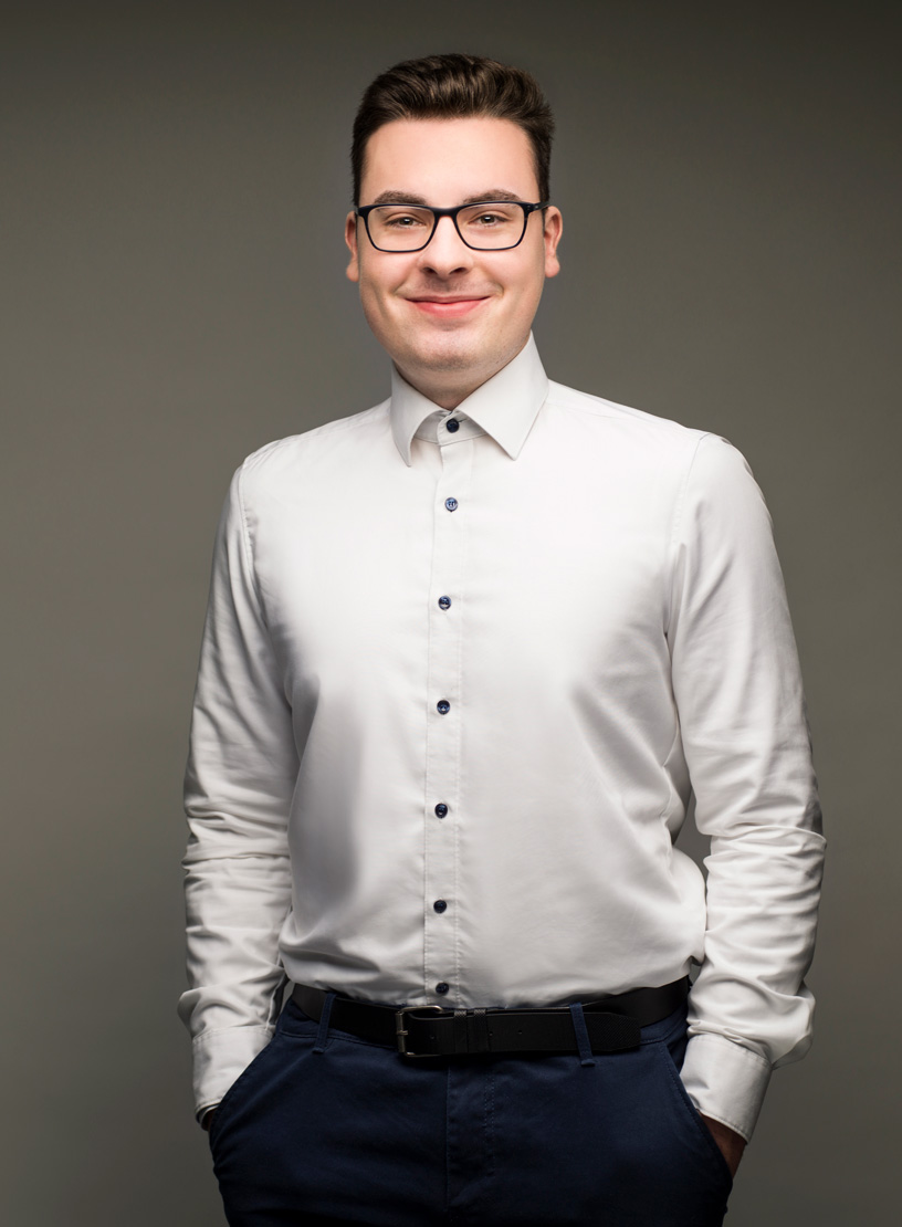Ein junger Mann mit Brille trägt ein weißes Hemd und dunkle Hose, steht mit verschränkten Armen vor einem grauen Hintergrund und lächelt freundlich.