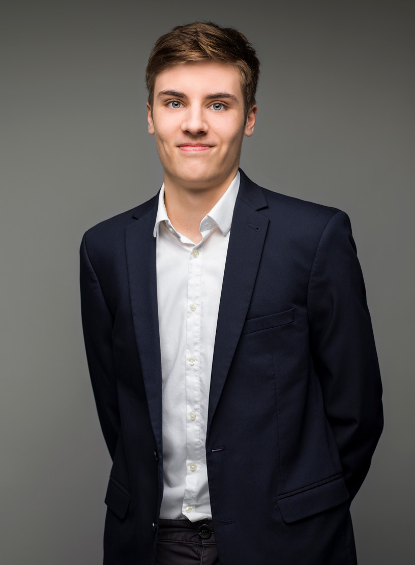 Junger Mann mit kurzer, blonder Haarfrisur trägt einen dunkelblauen Anzug und ein weißes Hemd, während er vor einem grauen Hintergrund steht.