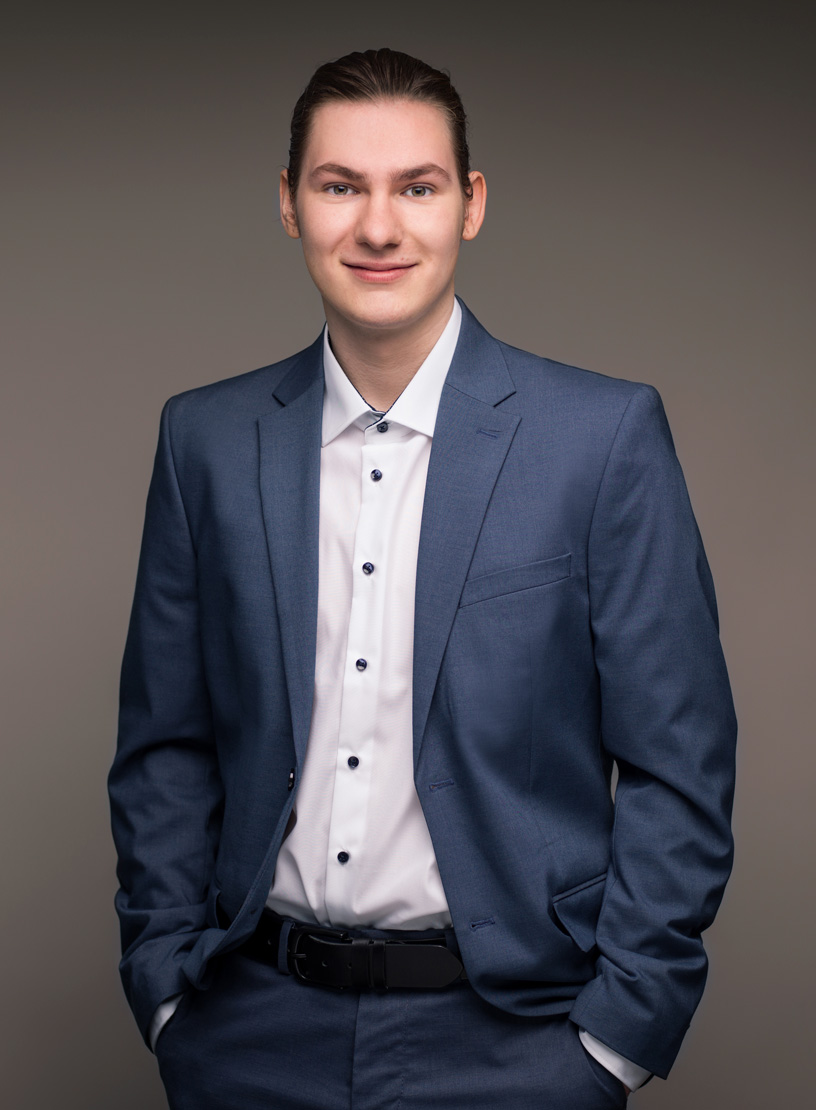 Ein junger Mann steht mit einem Lächeln vor einem grauen Hintergrund, trägt einen formellen blauen Anzug mit einer weißen Hemdbluse und hat die Hände entspannt in den Hosen gestützt.
