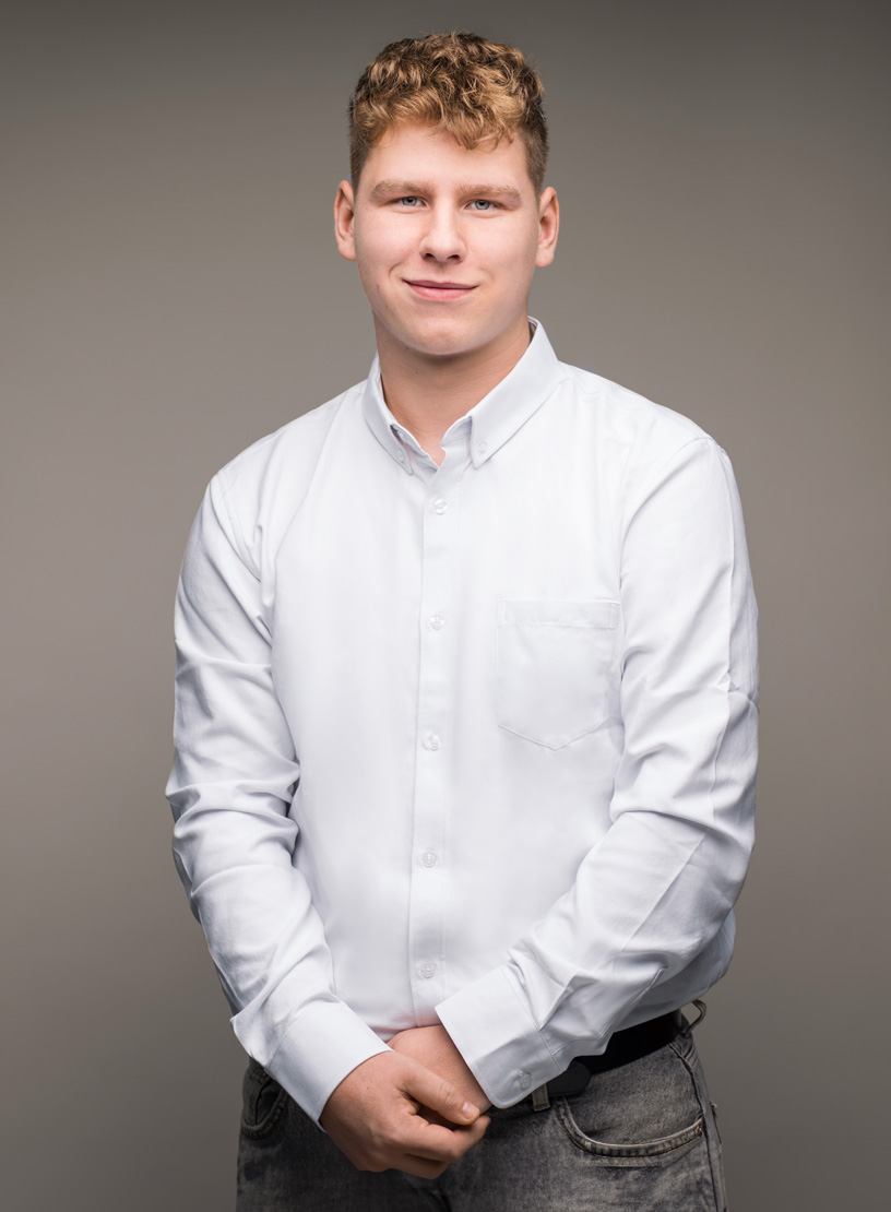Ein junger Mann mit kurzen, lockigen Haaren trägt ein weißes Hemd und schaut freundlich in die Kamera, während er seine Hände vor dem Bauch faltet. Der Hintergrund ist einheitlich grau.