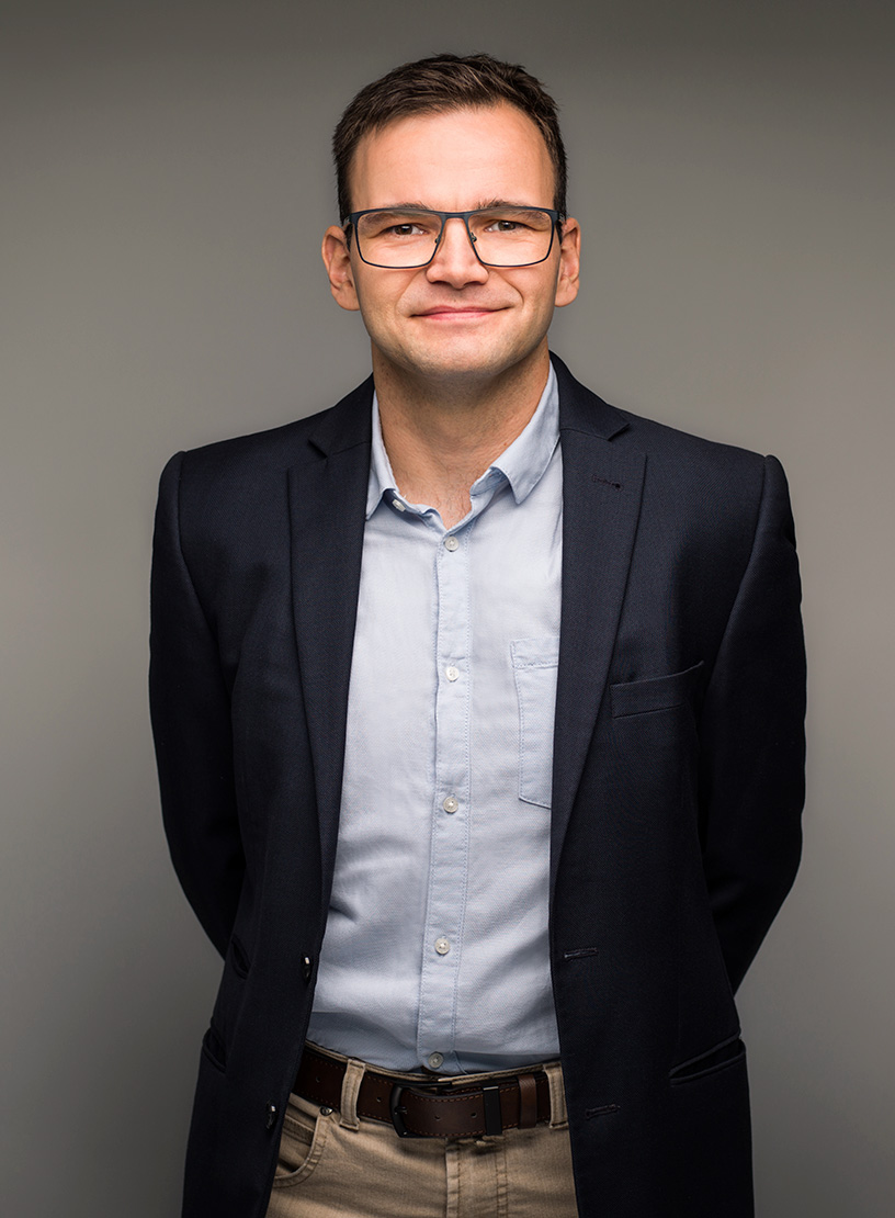 Ein freundlich lächelnder Mann trägt ein dunkelblaues Sakko über einem hellblauen Hemd und steht mit den Händen hinter dem Rücken vor einem neutralen Hintergrund.
