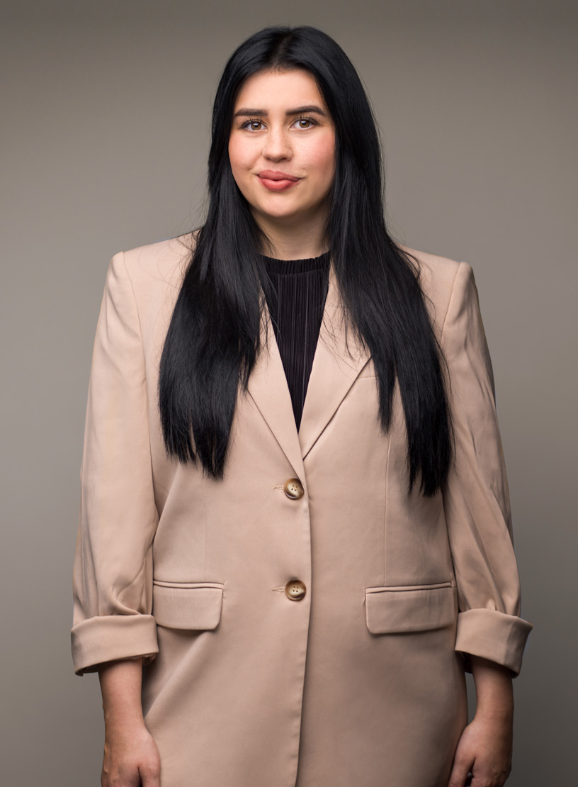 Eine junge Frau mit langen, schwarzen Haaren trägt einen beigen Blazer und schaut zur Kamera. Der Hintergrund ist einfarbig grau.