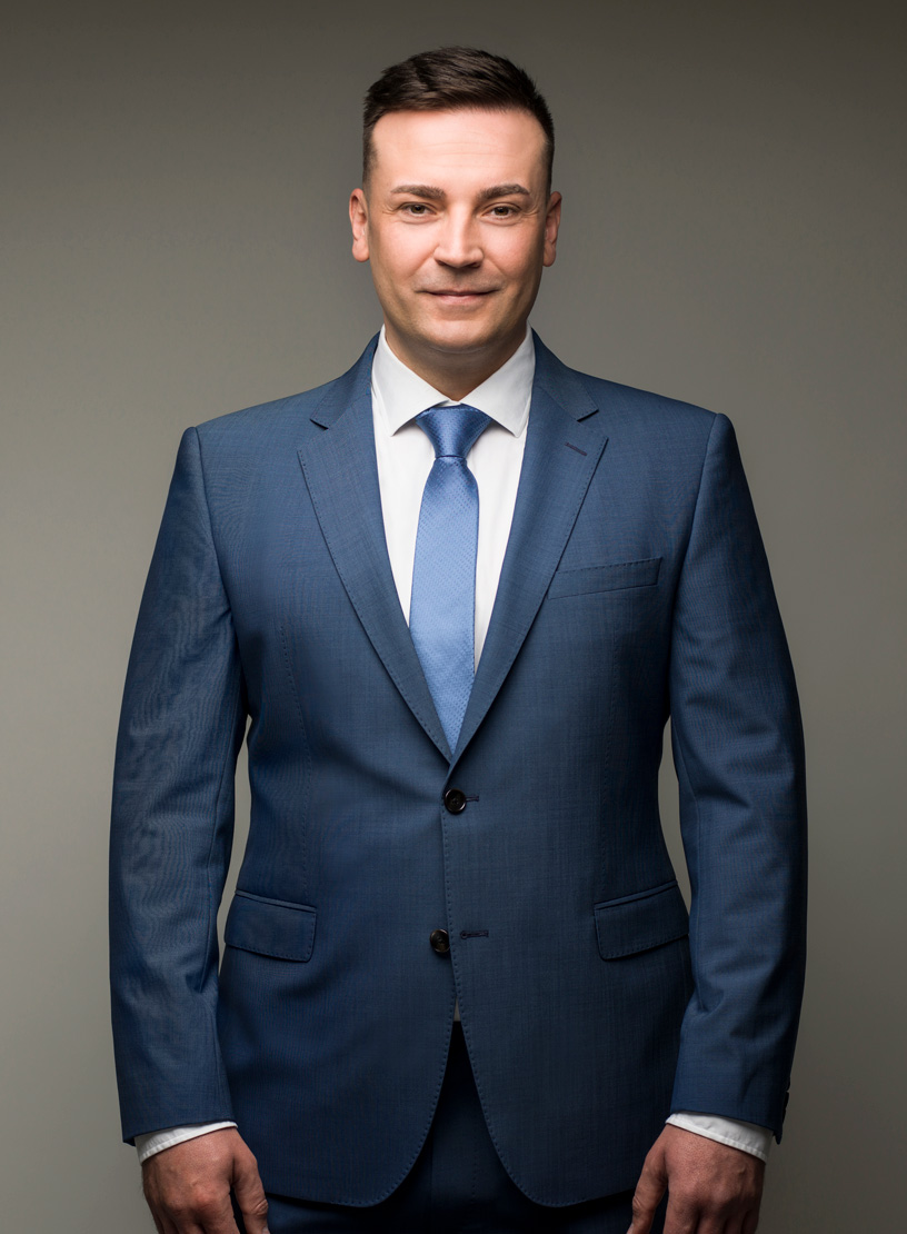 Ein Mann in einem eleganten blauen Anzug mit einem hellblauen Krawatte steht vor einem neutralen Hintergrund und schaut direkt in die Kamera.