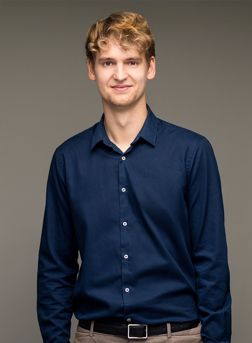 Ein junger Mann mit blonden lockigen Haaren trägt ein dunkelblaues Hemd und steht lächelnd vor einem grauen Hintergrund.