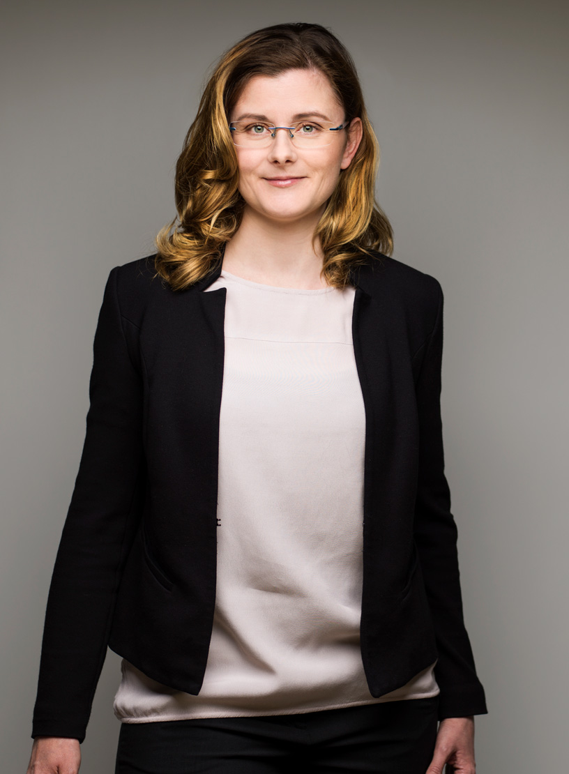 Eine selbstbewusste Frau mit langen, welligen Haaren trägt eine schwarze Jacke über einem hellen Oberteil und blickt direkt in die Kamera. Der Hintergrund ist neutral und hebt ihre Präsenz hervor.