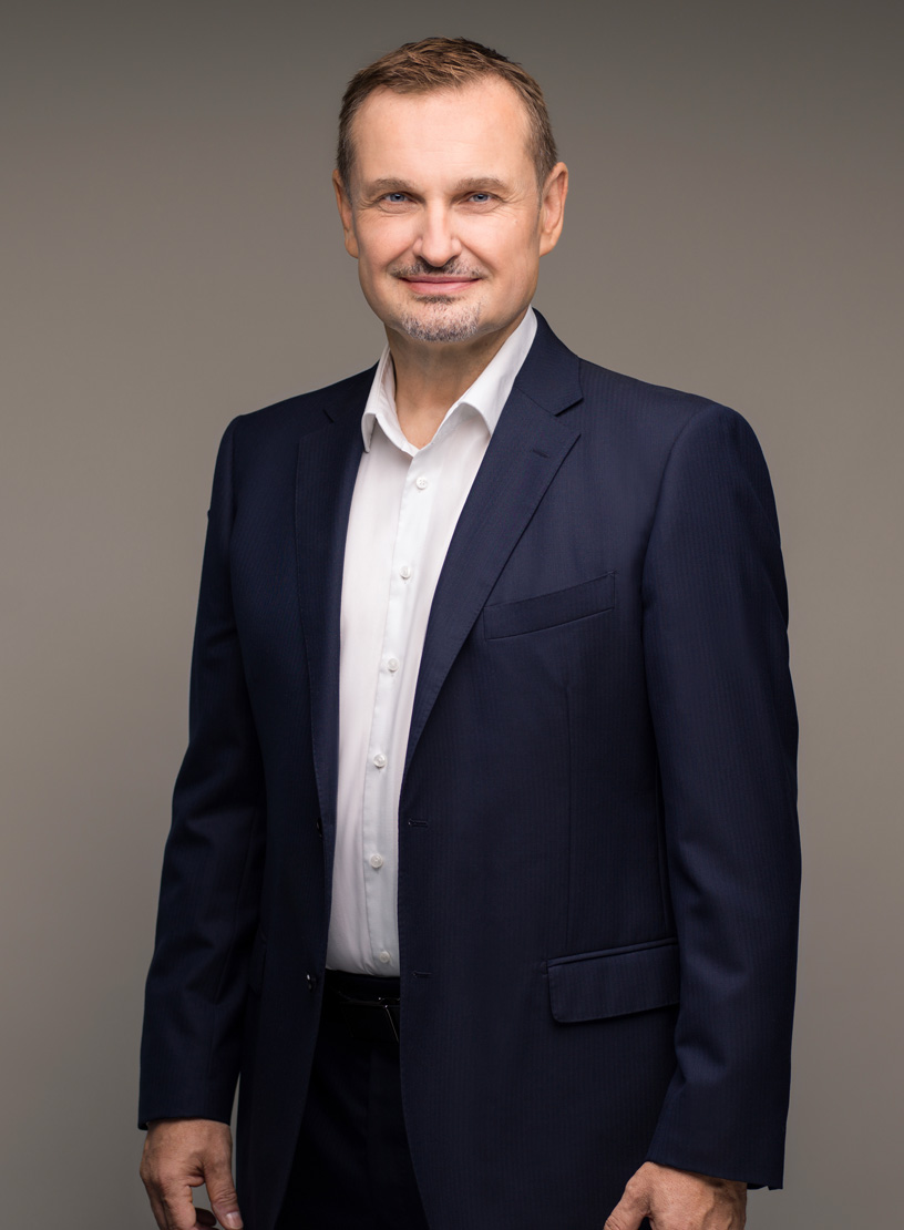 Ein mittelalterlicher Mann in einem dunklen Anzug und einem weißen Hemd steht aufrecht vor einem neutralen Hintergrund, lächelt freundlich und hat eine gepflegte Erscheinung.