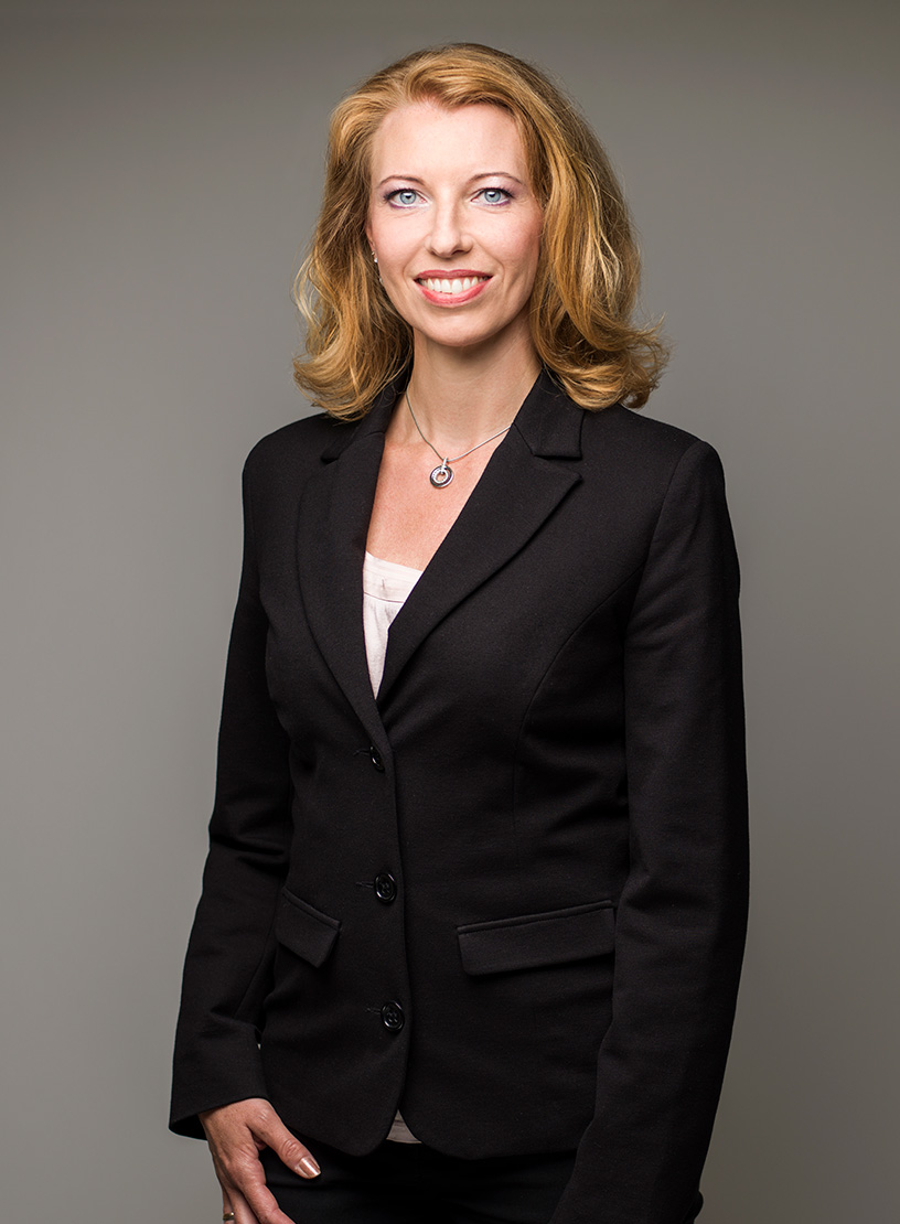 Eine Frau mit langen, blonden Haaren trägt einen schwarzen Blazer und steht in einer neutralen Pose vor einem einfarbigen Hintergrund. Sie lächelt freundlich in die Kamera.