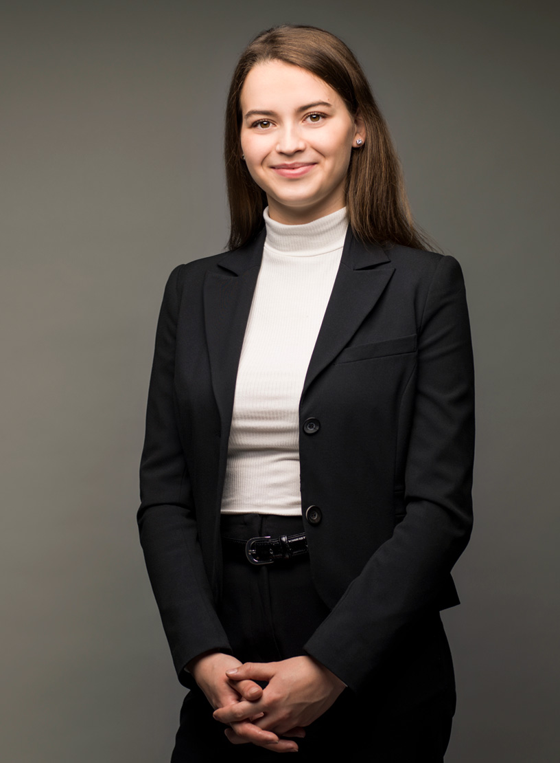 Eine junge Frau in einem schwarzen Blazer und weißem Rollkragenpullover steht lächelnd mit verschränkten Händen vor einem einfarbigen Hintergrund.