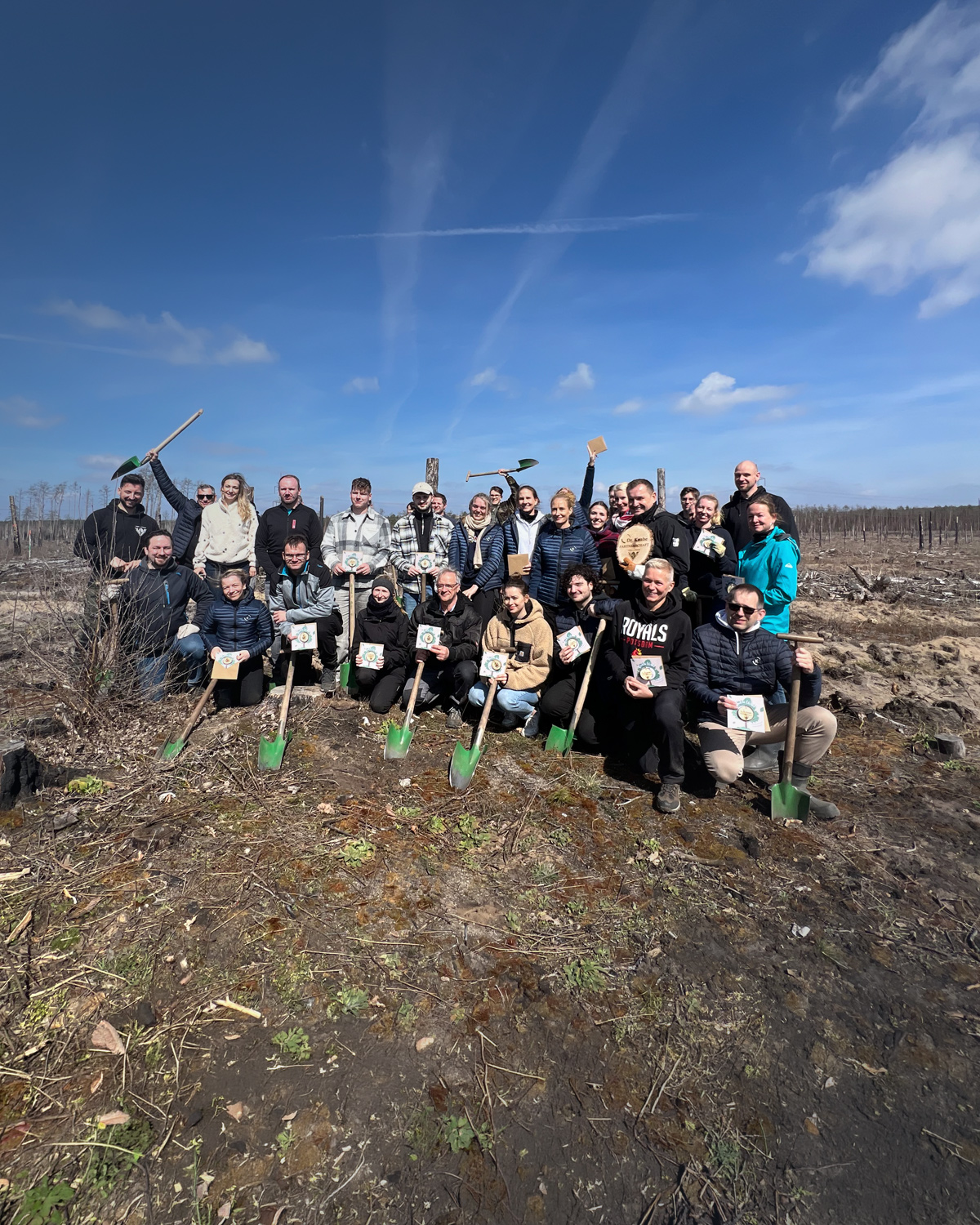 Gruppenshot von 24 Personen in einem kahlen Waldgebiet, alle halten Schaufeln und stehen in einer Pose des Feierns. Im Hintergrund sind teilweise abgeholzte Bäume und ein blauer Himmel mit wolkigen Abschnitten zu sehen.